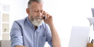 starszy pan z broda siedzący przed laptopem i rozmawiający przez telefon komórkowy