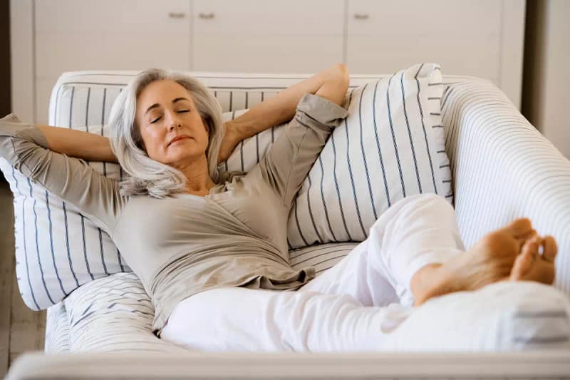Kobieta w średnim wieku leżąca na sofie z ułożonymi rękoma pod głową i zamkniętymi oczami z delikatnym uśmiechem na twarzy