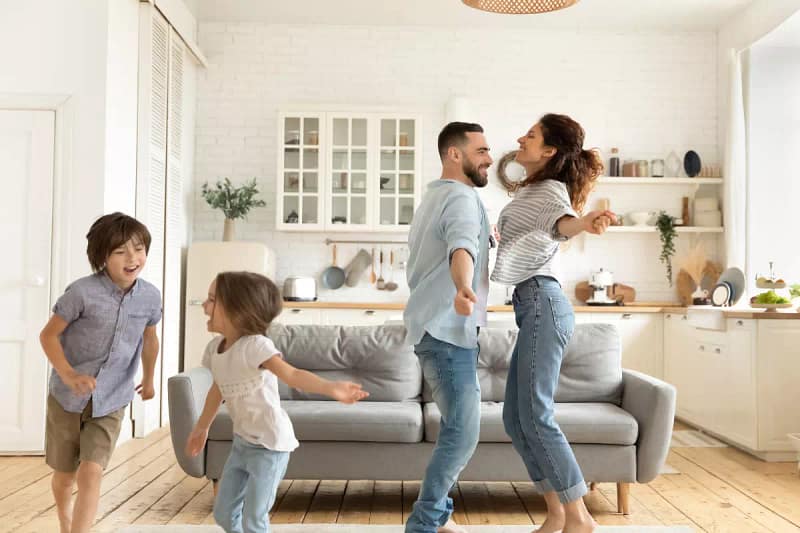 Szczęśliwa rodzina w pokoju. Rodzice tańczą a dwójka dzieci biega obok nich. W tle widoczna jest sofa oraz kuchnia